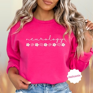 Puede incluir: Una sudadera rosa brillante con la palabra "neurology" en escritura blanca, acompa&ntilde;ada de flores y cerebros blancos. La sudadera es de cuello redondo y est&aacute; hecha de un material suave.