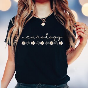 Puede incluir: Camiseta negra con la palabra "neurology" en cursiva. Debajo, seis flores blancas se alternan con ilustraciones de cerebros. La camiseta es de cuello redondo.