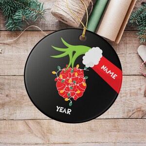 May include: A black Christmas ornament with a green hand holding a red heart-shaped design wrapped in Christmas lights. The ornament has the word "NAME" on a red stripe and the word "YEAR" at the bottom.