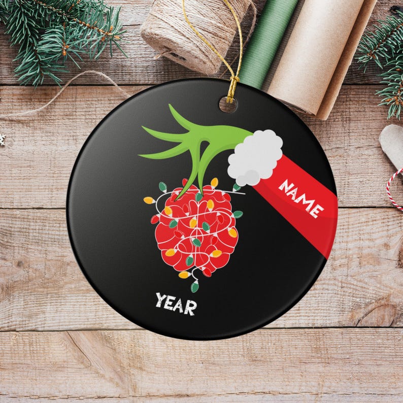 May include: A black Christmas ornament with a green hand holding a red heart-shaped design wrapped in Christmas lights. The ornament has the word "NAME" on a red stripe and the word "YEAR" at the bottom.