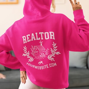 May include: A pink hoodie with the word "REALTOR" printed on the back, along with a house illustration and the text "YOURWEBSITE.COM".