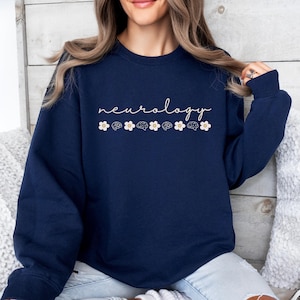 Puede incluir: Sudadera azul marino con la palabra "neurology" en cursiva. Debajo de la palabra, se alternan flores blancas e ilustraciones de cerebros. La sudadera es de cuello redondo.