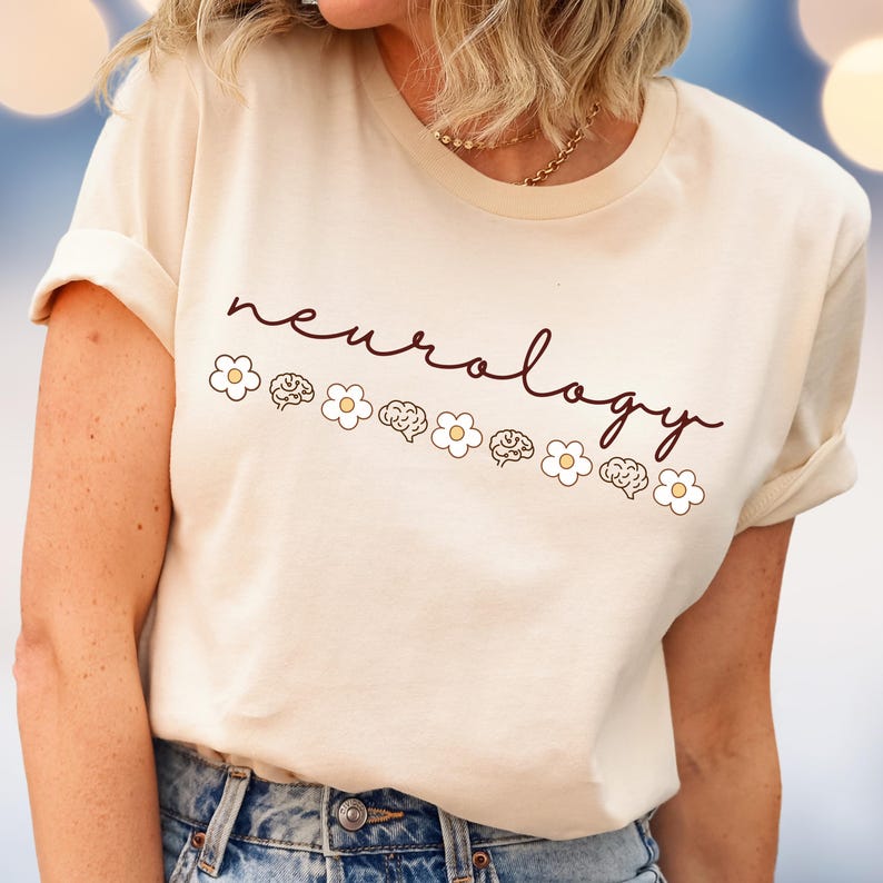 Puede incluir: Camiseta color crema con la palabra "neurology" en cursiva. Debajo, una fila de flores blancas y ilustraciones de cerebros. La camiseta tiene las mangas dobladas y se combina con unos vaqueros azules.