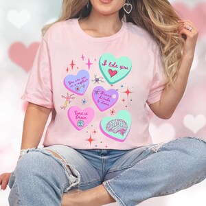May include: A light pink t-shirt featuring heart-shaped graphics with phrases like "I lobe you" and "Brains, Scrubs & Love." The design includes a brain illustration and star accents. The shirt is paired with blue jeans.