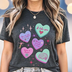 May include: Dark gray t-shirt featuring five heart-shaped designs with brain-related graphics and text. The hearts are in shades of pink and teal, with phrases like "I love you" and "time is brain".