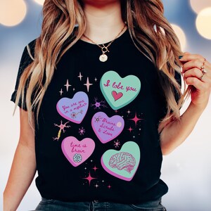 May include: Black t-shirt featuring five heart-shaped designs with science-themed text and illustrations. The hearts are various colors, including teal, purple, and pink, with phrases like "I lobe you" and "Brains Scrubs & Love."