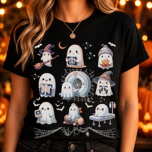 May include: A black t-shirt featuring a graphic print of multiple cartoon ghosts in various medical and Halloween-themed scenarios, including ghosts with X-rays, a CT scanner, a hospital bed, and witch hats, surrounded by small stars and spider webs.