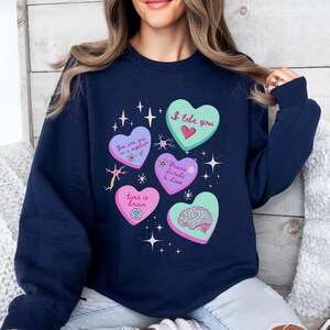May include: Navy blue sweatshirt with heart-shaped graphics and text. The hearts feature phrases like "I lobe you," "You are one in a myelin," and "Time is brain." The design includes brain illustrations and star accents.