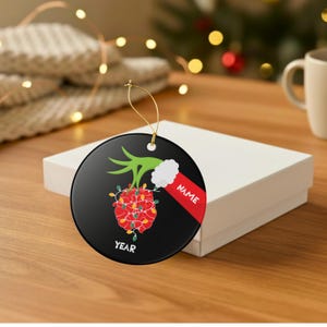 May include: A black Christmas ornament with a green hand holding a red heart wrapped in Christmas lights. A red sleeve with the word "NAME" and the word "YEAR" are also on the ornament.