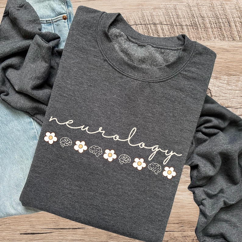 Puede incluir: Sudadera gris oscuro con la palabra "neurology" en escritura cursiva, acompa&ntilde;ada de margaritas y gr&aacute;ficos de cerebro. La sudadera est&aacute; doblada sobre una superficie de madera, con unos vaqueros azules visibles.