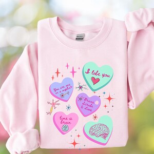 May include: A light pink sweatshirt with several heart-shaped graphics. The hearts feature phrases like "I lobe you" and "Brains Scrubs & Love", along with brain and neuron illustrations. The Gildan brand label is visible.