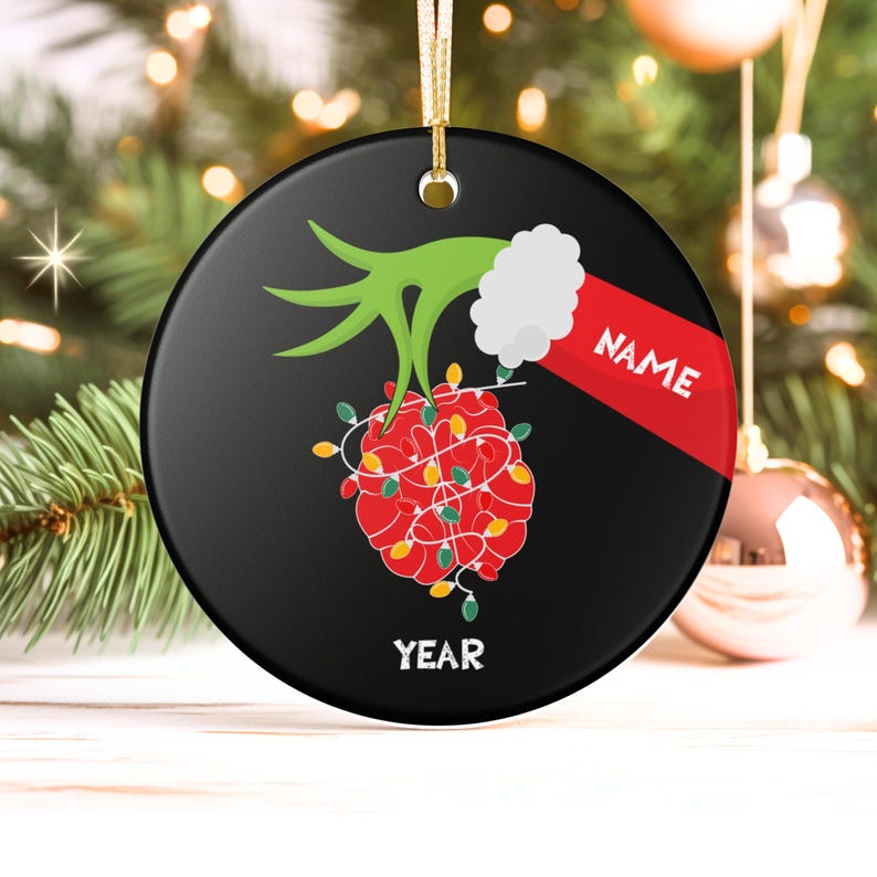 May include: Black round Christmas ornament with a green hand holding a red ball wrapped in Christmas lights. The ornament has the word "NAME" on a red banner and the word "YEAR" at the bottom.