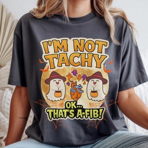 May include: Dark gray t-shirt with a Halloween-themed graphic. The design features two cartoon ghosts wearing hats and holding pumpkins, with the text "I'M NOT TACHY OK... THAT'S A-FIB!" above and below a full moon.