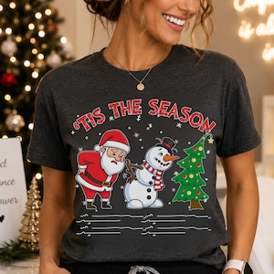 May include: A dark gray short-sleeve t-shirt featuring a graphic print of Santa Claus and a snowman both holding their lower backs in pain. Above them, the text reads 'TIS THE SEASON in red letters, with a green Christmas tree to the right.
