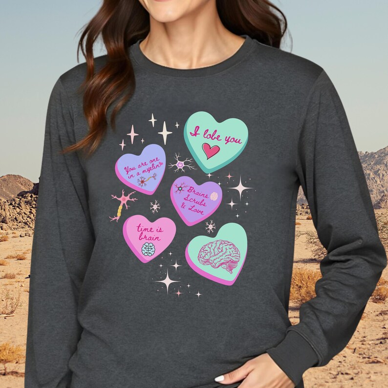 May include: Dark gray long-sleeve shirt featuring heart-shaped graphics with text and illustrations. The hearts are various colors, including blue, pink, and green, with phrases like "I lobe you" and "Brains Scrubs & Love". The shirt has a crew neck.