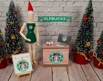 May include: A miniature elf barista stands behind a counter with a sign that reads "Elfbucks Coffee". The counter is decorated with a green and white coffee cup logo. There are small Christmas trees and a sign that says "It's a coffee thing!" in front of the counter.