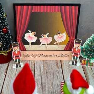 Könnte beinhalten: Eine festliche Szene mit einem roten und goldenen Bühnenhintergrund, auf dem drei Ballerinas in rosa Tutus zu sehen sind. Die Bühne ist mit "The Elf Nutcracker Ballet" beschriftet. Zwei Nussknackerfiguren stehen zu beiden Seiten der Bühne, und im Vordergrund stehen zwei Weihnachtsbäume.