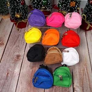 May include: A collection of ten mini backpacks in various colours, including purple, pink, yellow, orange, red, black, brown, white, blue, and green. The backpacks are arranged in two rows on a wooden surface.