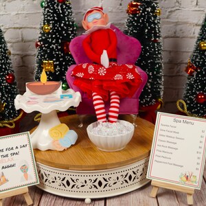 以下が含まれることがあります： 赤い白のドレスを着てピンクのアイマスクをしたクリスマスエルフのドールが、ピンクのベルベットの椅子に座ってフットバスをしています。エルフは、「スパで一日過ごしましょう！リラックス...Ahhh！」と書かれた看板と、様々なサービスの価格が書かれたスパメニューのあるスパにいます。