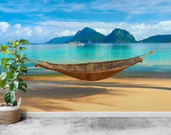 May include: A hammock hangs between two palm trees on a tropical beach. The hammock is made of woven brown material and is suspended by ropes. The beach is sandy and the water is turquoise blue. In the background, there are lush green mountains and a small boat.