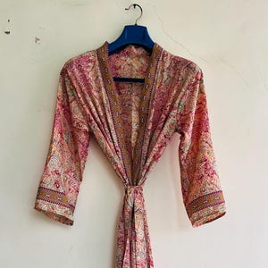 May include: A pink paisley patterned robe with a matching tie, hanging on a blue hanger. The robe has three-quarter sleeves and a decorative border along the edges. The fabric features a mix of pink, yellow, and green hues.