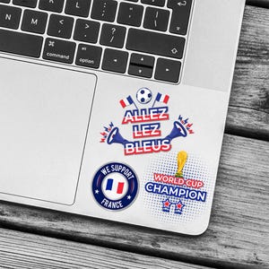 May include: A set of stickers featuring French football designs. The stickers include the text "ALLEZ LEZ BLEUS", a soccer ball, the French flag, and the words "WORLD CUP CHAMPION". The stickers are on a laptop.
