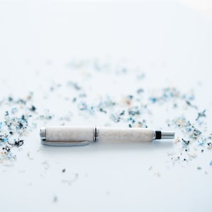 May include: A white fountain pen with silver accents and a black ink cartridge lies on a white surface with scattered pencil shavings.