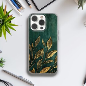 Puede incluir: Una funda transparente para teléfono con un fondo verde oscuro y un diseño de hojas doradas. La funda tiene un borde transparente y una abertura para la cámara. Las hojas están detalladas con contornos dorados y dispuestas en un patrón botánico.