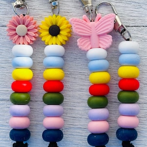 May include: Four colourful beaded lanyards with flower and butterfly charms. Each lanyard features a series of round beads in various colours, including white, blue, yellow, red, green, purple, pink, and navy. The charms include a pink flower, a yellow sunflower, and a pink butterfly.