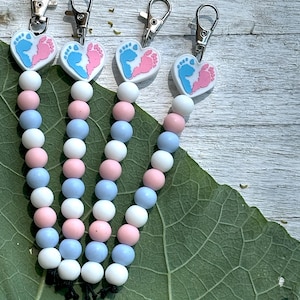 May include: Four white heart-shaped charms with blue and pink footprints, attached to a string of alternating pink and blue beads. Each charm has a silver key ring attached.