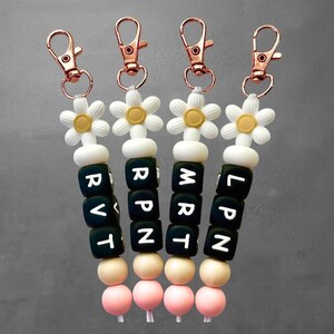 Puede incluir: Cuatro llaveros con dijes de flores de margarita y cierres de oro rosa. Cada llavero tiene un conjunto diferente de cuentas de letras que deletrean RVT, RPN, MRT y LPN. Las cuentas están ensartadas con cuentas blancas, beige y rosas.