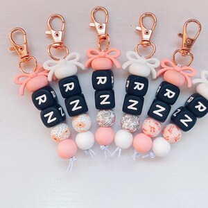 Peut inclure: Ensemble de six porte-clés avec des perles roses, blanches et noires. Chaque porte-clés est orné d'un charme avec un nœud et les lettres "RN" en noir.