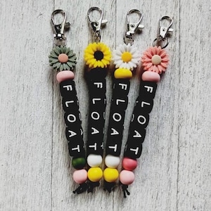 May include: Four keychains with black beads spelling out the word "FLOAT" in white letters. Each keychain has a different coloured flower charm: green, yellow, white, and pink.