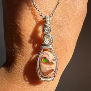 May include: A handcrafted silver pendant necklace featuring a wire-wrapped opal gemstone. The opal displays a mix of colors, including green, red, and orange. The pendant hangs from a delicate silver chain, showcasing the unique stone.
