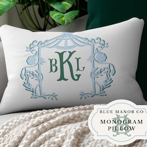 May include: A white pillow with a blue and green monogram design. The monogram is surrounded by a decorative floral frame. The pillow is labeled "Blue Manor Co. Monogram Pillow".