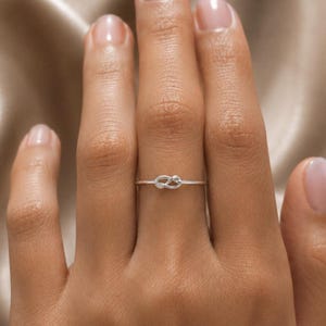 Peut inclure: Une fine bague en argent poli présentant un petit nœud central. L'anneau est mince et circulaire, avec une finition métallique qui reflète la lumière. L'élément en forme de nœud est placé au centre de la bague, créant une structure en boucle continue sur l'anneau.