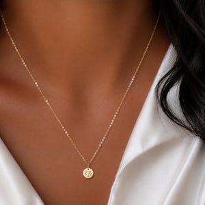 May include: A gold-toned necklace featuring a delicate chain and a small, circular pendant. The pendant is engraved with the letters KB in a cursive script font. The necklace rests against a white fabric garment, showcasing a simple and refined design.