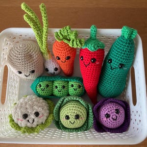 May include: A collection of crocheted plush vegetables with embroidered smiling faces, including a potato, spring onion, carrot, chilli pepper, cucumber, pea pod, cauliflower, cabbage, and purple cabbage, all arranged inside a white plastic basket.