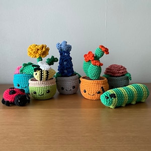 May include: A collection of seven crocheted potted plants, including a dandelion, a cactus, a blue flower, a bee, and a caterpillar. The pots are all different colours and have happy faces.