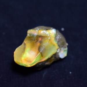 May include: A rough-cut opal gemstone with a mix of colors, including yellow, green, and hints of orange. The stone has a natural, irregular shape and a slightly translucent appearance. The opal is set against a dark background.