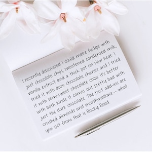 May include: A handwritten note on white paper with a silver pen resting on top. The note describes a recipe for fudge with chocolate chips, condensed milk, vanilla extract, and chocolate chunks. The note mentions that the best add-ins are crushed almonds and marshmallows.
