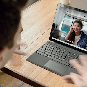 May include: A person is on a video call on a black laptop with a keyboard and trackpad. The person on the screen is smiling and has long dark hair. The laptop is on a wooden table.