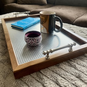 May include: A rectangular wooden serving tray with a woven metal surface, silver handles, and a dark brown frame. The tray holds a blue book, a patterned bowl, and a black and gold mug. The tray is on a soft, textured surface.