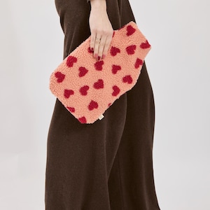 May include: A peach-coloured, rectangular clutch bag featuring a repeating pattern of red hearts. The bag is made of a fuzzy, textured material. The person is wearing brown trousers and a brown top.