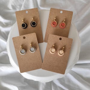 Könnte beinhalten: Vier Paar handgefertigte Ohrringe, präsentiert auf braunen Karten. Jedes Paar hat einen kleinen, farbigen Kreis mit einem goldfarbenen Ring und Stecker. Zu den Farben gehören Schwarz, Rot-Orange, Grau und Creme. Die Ohrringe werden auf einem weißen Teller mit gewelltem Rand präsentiert.