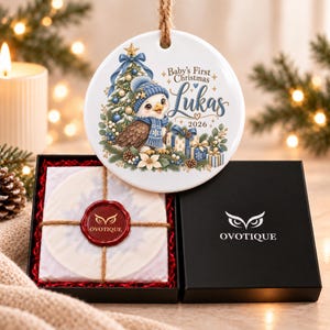 Peut inclure: Une décoration ronde en céramique blanche ornée d'un hibou portant un bonnet et une écharpe bleus près d'un sapin et de cadeaux. Le texte indique Baby's First Christmas Lukas 2026. L'objet est présenté dans une boîte noire avec le logo Ovotique.