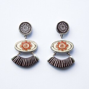 Puede incluir: Un par de pendientes colgantes con acabado plateado. Los pendientes presentan tres niveles de colgantes circulares y ovalados con diseños geométricos en blanco y negro y acentos naranjas.