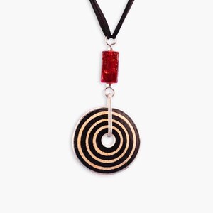 May include: A black and white circular pendant with concentric circles hangs from a black cord. A red rectangular bead is above the pendant.