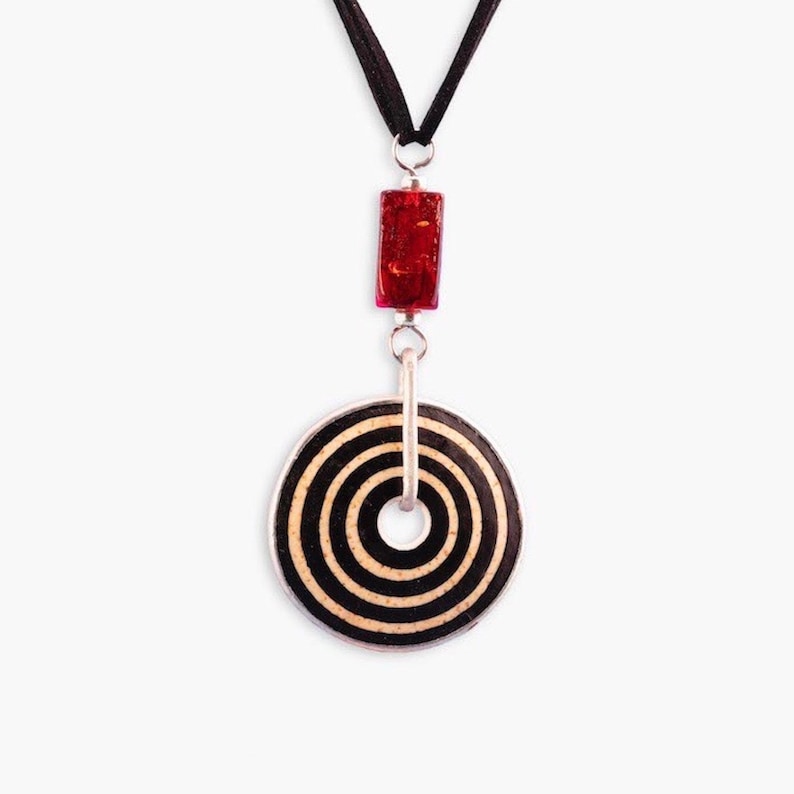 May include: A black and white circular pendant with concentric circles hangs from a black cord. A red rectangular bead is above the pendant.