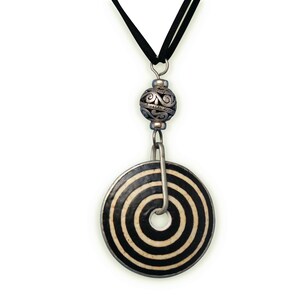 May include: A black and white ceramic pendant with a spiral design hangs from a black cord. The pendant is attached to a silver bead with a decorative design.